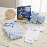 A baby gift set arranged on a cream blanket, including a light blue hooded onesie, matching pants, a white short-sleeve bodysuit with animal graphics, a blue knitted teddy bear, a blue textured blanket, a book titled 'I Love You in the Moon and Back', and a round wooden 'Hello World' card.