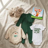 A baby clothing flat lay on a beige linen surface, including a dark green footed sleeper, a cream long-sleeve bodysuit with a golf graphic and text, a white bodysuit with a golf-themed slogan, a teddy bear on a knit blanket, and a children's book titled Golf Stars.