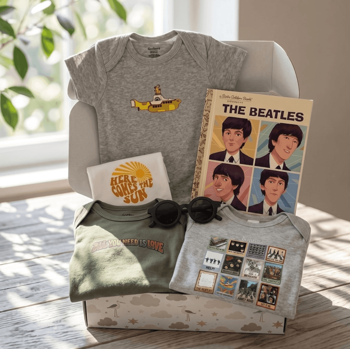 A curated Beatles-themed baby gift set arranged in a white box on a sunlit wooden floor. The set includes a heather gray baby bodysuit with a yellow submarine graphic, a white T-shirt with a yellow 'Here Comes the Sun' print, an olive green long-sleeve shirt with 'The Power of Love Is Love' text, a light gray T-shirt with retro album-cover graphics, a book titled 'The Beatles', and black sunglasses.