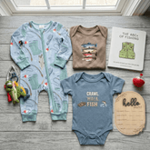A baby clothing flat lay on a gray wood floor featuring a light blue zip-up onesie with a green turtle print, a brown short-sleeve shirt with a stacked fish graphic, a slate blue bodysuit with 'CRAWL WALK FISH' text, a book titled 'THE ABCS OF FISHING', a wooden 'hello world' sign, and a small red apple.