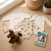 Baby clothing and toys on a wooden floor with a book and plush toy.