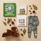 A styled flat-lay of baby apparel and a children's book on a light wood surface, surrounded by pine branches, acorns, and a teddy bear. The set includes a green book titled 'WINDOWS TO THE NATIONAL PARKS,' a cream baby bodysuit with 'FUTURE PARK RANGER' text, a white baby top with landscape prints, and a teal footed sleeper with outdoor animal and scenery prints.