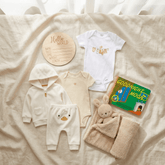 A baby outfit and accessories set arranged on a cream fabric background, including a white bodysuit with a bunny graphic, a cream hooded jacket, cream pants with a duck face, a cream striped bodysuit, a round wooden name plaque, a knitted bunny toy, and a green 'Goodnight Moon' book.