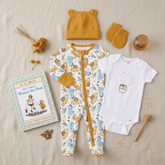 A styled flat-lay of a baby clothing set on a beige fabric background, including a mustard yellow beanie, matching mittens, a white short-sleeve bodysuit with a honey jar graphic, and a one-piece footed sleeper with a multicolor animal print and mustard trim. A children's book, honey dipper, bee ornament, oats, and dried flowers are arranged around the outfit.