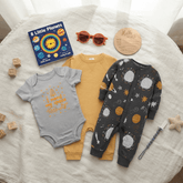 Three baby onesies arranged on a white fabric surface with playful props: a light gray short-sleeve bodysuit with orange space-themed text, a mustard yellow long-sleeve bodysuit, and a dark gray one-piece with a white and yellow space print. A book, sunglasses, wooden blocks, and a round sign are also visible.