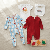 A baby holiday-themed clothing set laid flat on a light beige fabric background, including a light blue one-piece with Santa Claus face prints, a white short-sleeve bodysuit with a Santa face graphic, a white bib reading 'My First Christmas,' a red footed sleeper, and a small wooden gingerbread cookie puzzle.