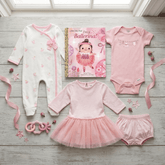 A coordinated baby gift set arranged on a light wood floor near a window: a white footless onesie with pink trim and floral print, a pink short-sleeve bodysuit, a pink tutu dress, matching pink bloomers, a ballet-themed book, and pink ribbons and toy accessories.