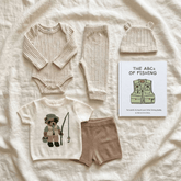 A baby clothing flat lay on a cream fabric background, including a cream cable-knit long-sleeve bodysuit, matching cream knit pants, a cream knit hat, a cream graphic T-shirt with a teddy bear fishing design, tan knit shorts, and a children's book titled 'THE ABCs OF FISHING'.