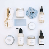 A flat-lay arrangement of lavender-themed bath and body products from Lavender Meadows Co. on a white background, including a jar of body wash, a bottle of shower foam, a pump bottle of coconut oil, a jar of body butter, a small tin, a blue-and-white scrunchie, and dried lavender and wheat stems.