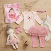 A coordinated baby ballerina outfit set arranged on a light wood surface, including a pale pink long-sleeve bodysuit with a ballerina print, a pink ruffled tutu skirt, pink leggings with white dots, pink booties, a pink bow headband, a pink bow headband, a doll, a book titled 'I'm a Ballerina!', and small dried flowers.