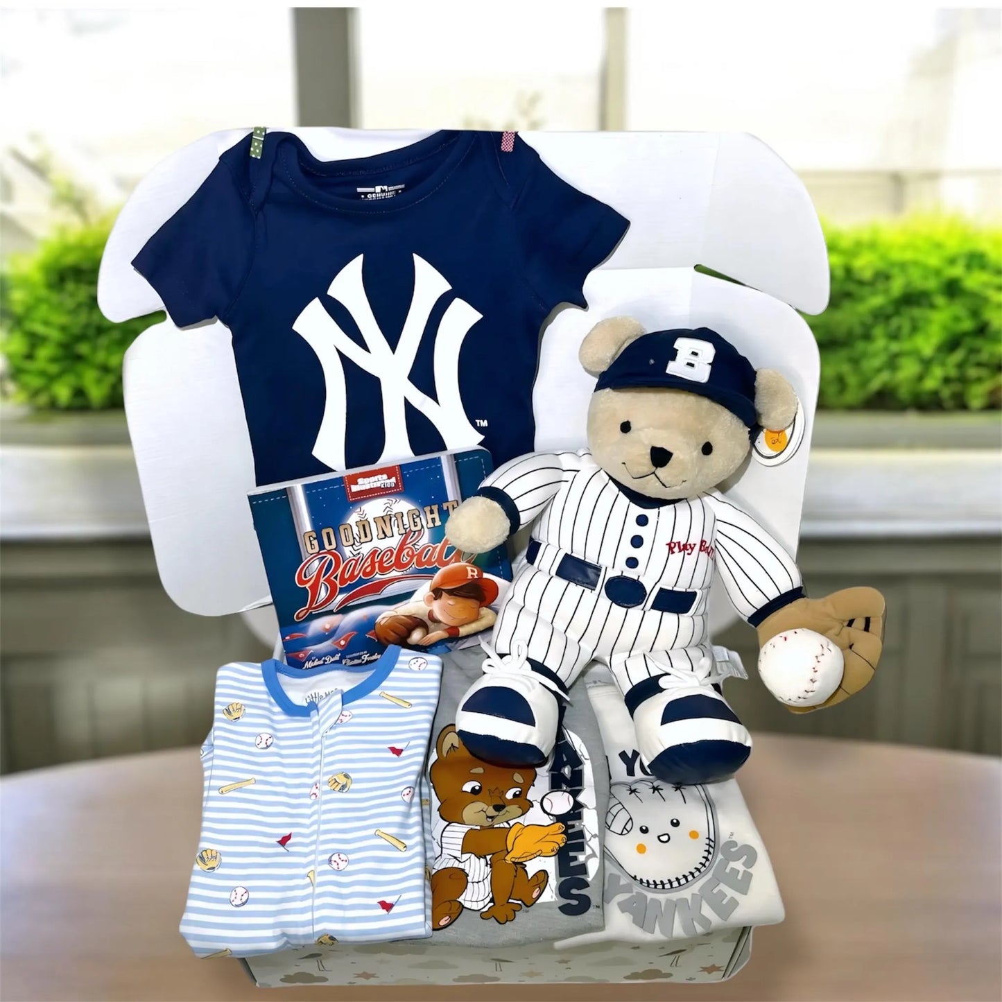 Baby gift set with Yankees-themed items including a onesie, teddy bear, and bib on a white stand.