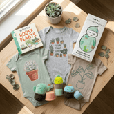 A styled flat-lay of baby gift items on a light wood surface: a gray bodysuit with a cactus and plant print, a sage green bodysuit with a succulent print, a beige bodysuit with a plant outline print, a book titled 'My First Book: House Plants,' a boxed knit set labeled 'little sleepies,' and small plush plant toys.