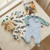 Two baby onesies inspired by 'The Child' from 'Star Wars' on a wooden surface with a 'Star Wars' book.