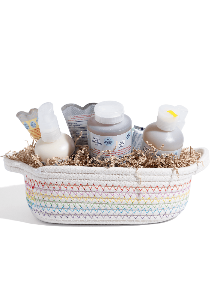 A baby gift basket set in a white woven basket with pastel rainbow stripes, filled with baby care products including a cream-colored bottle, a gray bottle, a small tube, and a heart-shaped item, all arranged on brown paper shred against a white background.