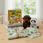 A baby gift set arranged on a wooden table in front of a softly blurred window. The set includes a folded light green baby blanket or swaddle printed with woodland animals, a matching folded cloth piece, a plush brown bear toy, a plush raccoon toy, a gray folded fabric item, a small wooden bead toy, and a children's book titled 'All Aboard! Wildlife Adventures in Parks'.