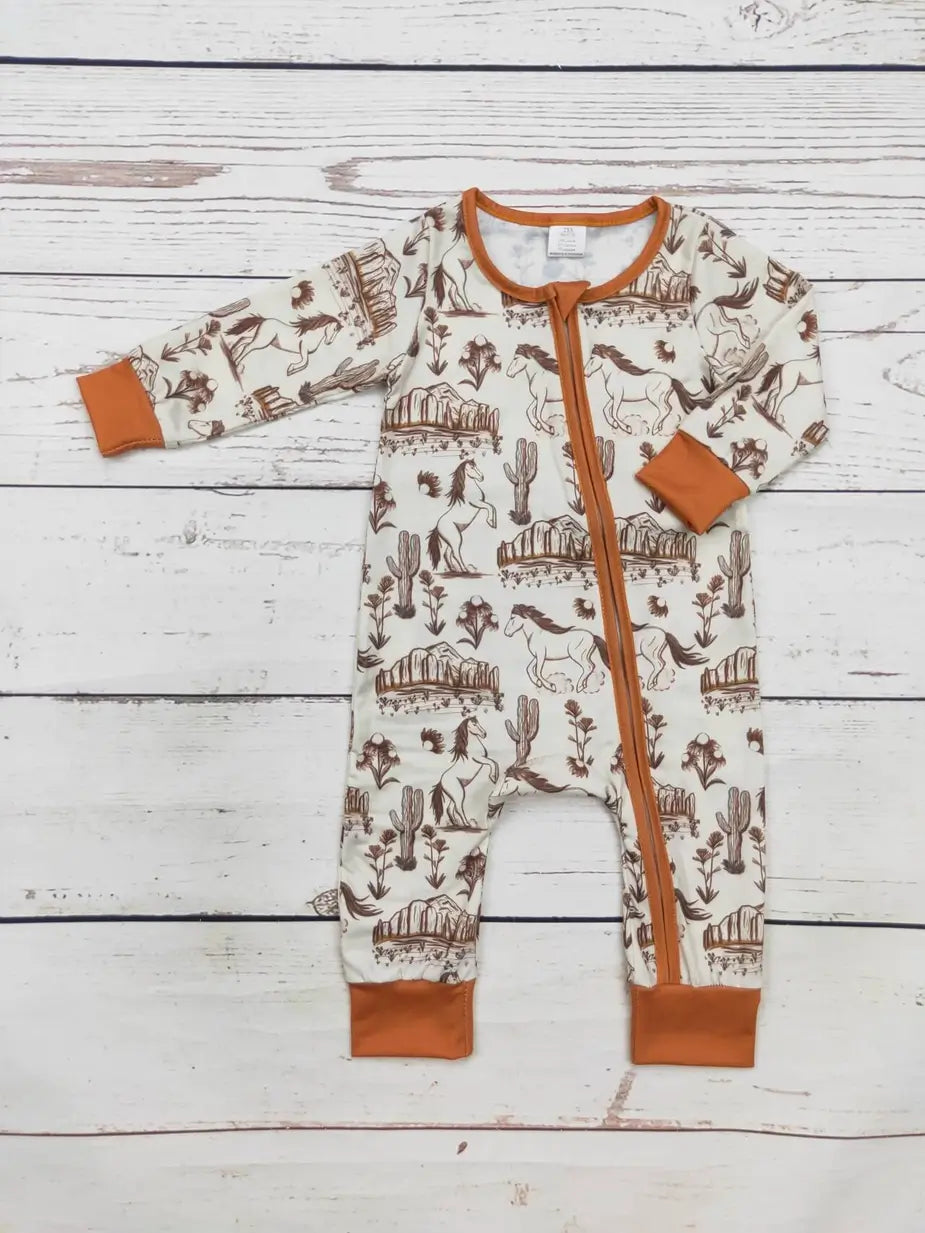 A baby long-sleeve footless sleeper laid flat on a white wood plank background. The sleeper has an off-white base with a brown western-themed print of horses, cacti, and fences, and features orange trim along the zipper, cuffs, and collar.