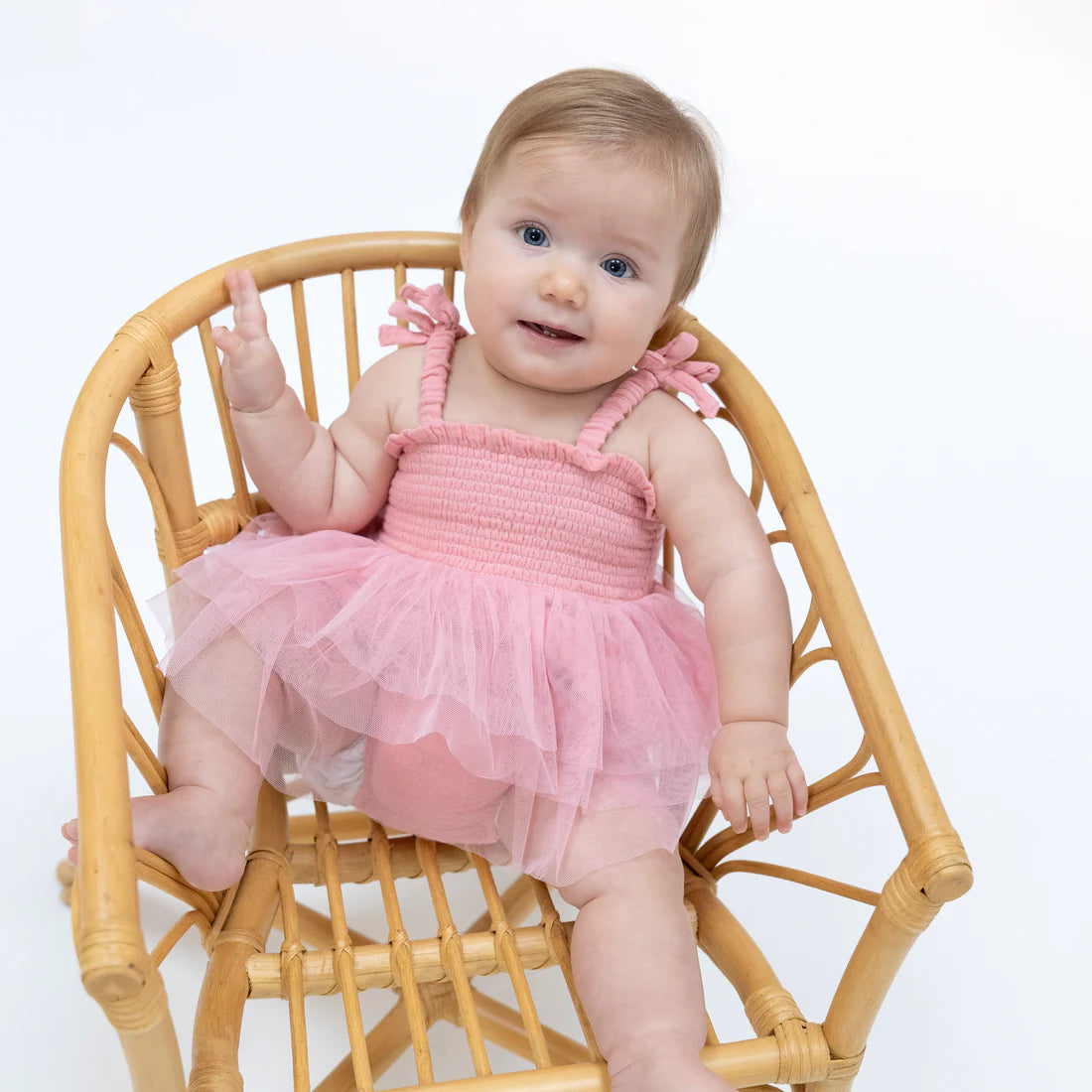 Baby in a pink dress sitting on a wicker chair against a white background
