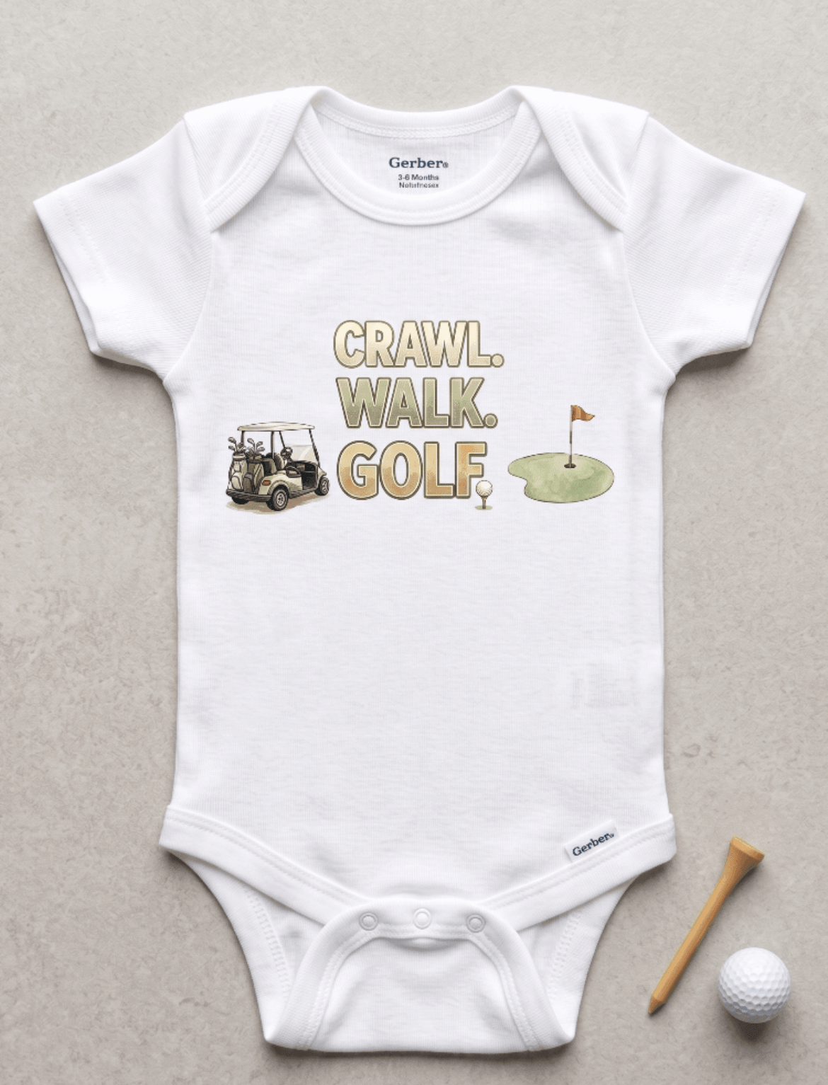 A white short-sleeve baby bodysuit laid flat on a light gray surface, featuring a golf-themed graphic with the words 'CRAWL, WALK, GOLF' and small illustrations of a golf cart, golf ball, and green hole flag. A wooden tee and golf ball are placed beside the garment.