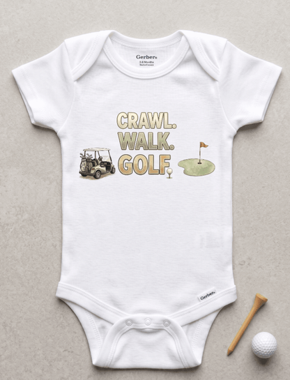 A white short-sleeve baby bodysuit laid flat on a light gray surface, featuring a golf-themed graphic with the words 'CRAWL, WALK, GOLF' and small illustrations of a golf cart, golf ball, and green hole flag. A wooden tee and golf ball are placed beside the garment.
