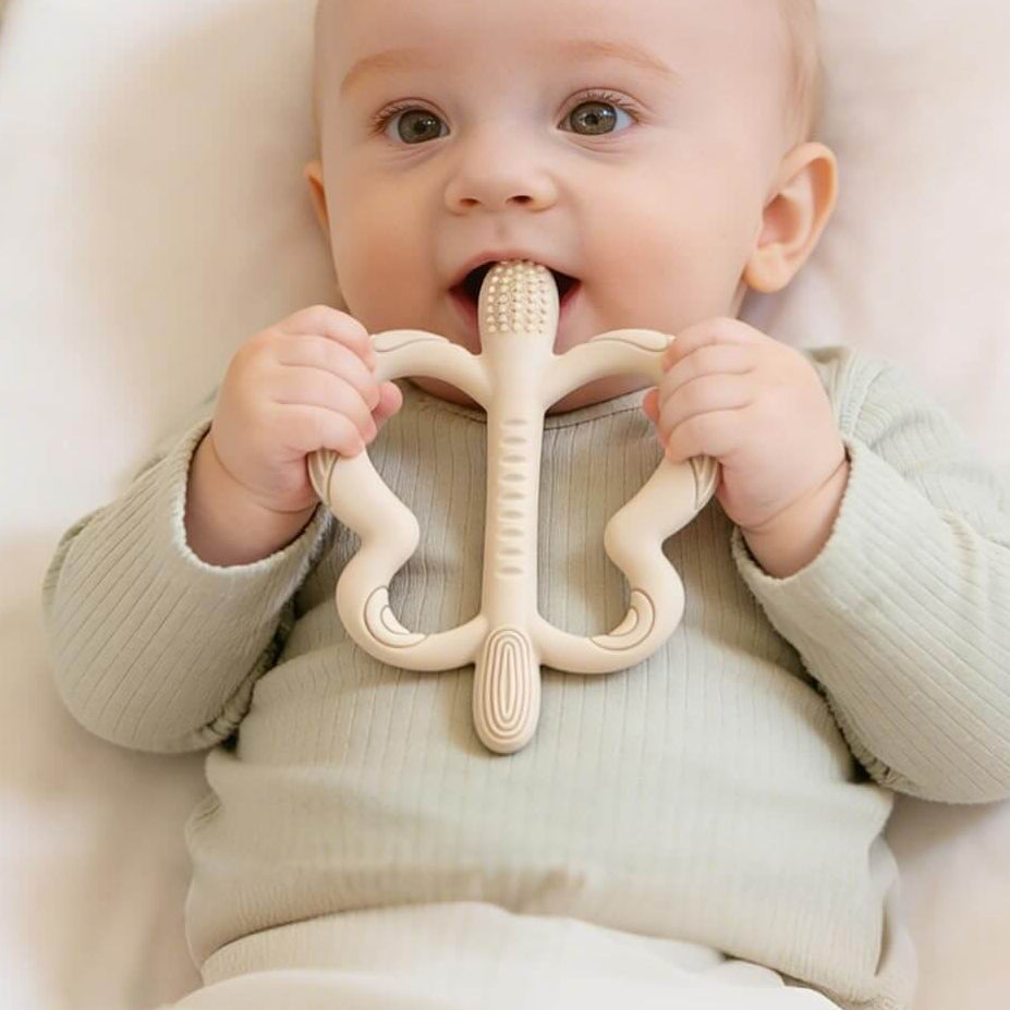 A baby wearing a light sage green ribbed onesie lies in a white crib, holding a natural beige wooden teether with curved arms and a central handle. Blue text at the top reads 'Safe for Baby's Delicate Mouth'.