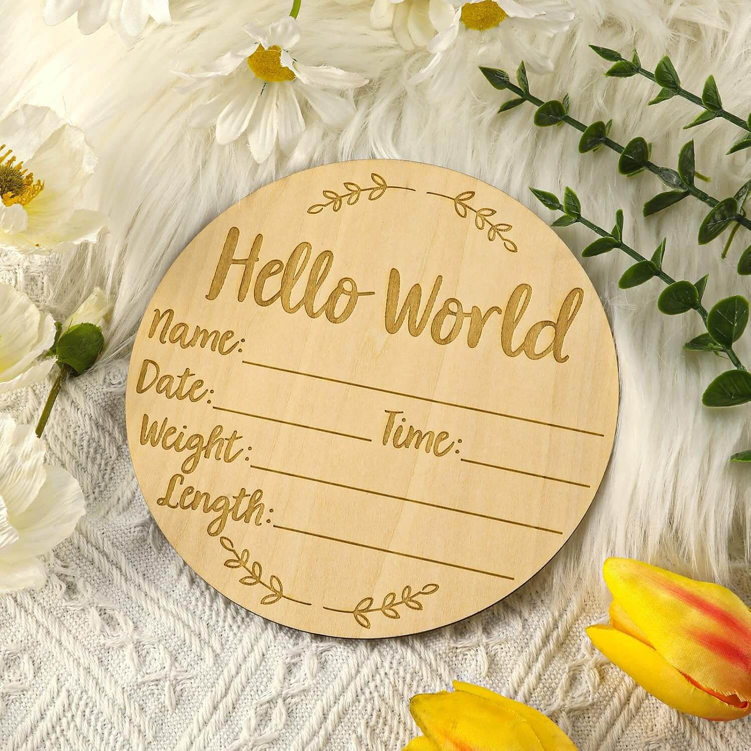 A round natural wood engraved baby milestone plaque reading 'Hello World' with lines for Name, Date, Weight, and Time, styled on a white textured blanket with white flowers, green leaves, and yellow tulips.