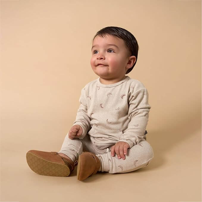 A baby sitting on a warm beige studio background, wearing a light cream long-sleeve romper with a small crescent moon print and tan booties.