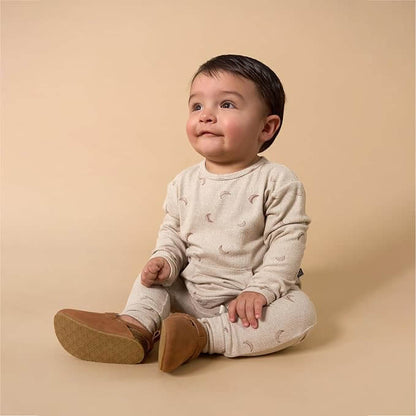 A baby sitting on a warm beige studio background, wearing a light cream long-sleeve romper with a small crescent moon print and tan booties.