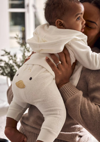 A baby wearing a cream-colored fleece hoodie and matching pants with a duck face graphic, held by an adult in a brown knit sweater in a cozy indoor setting.