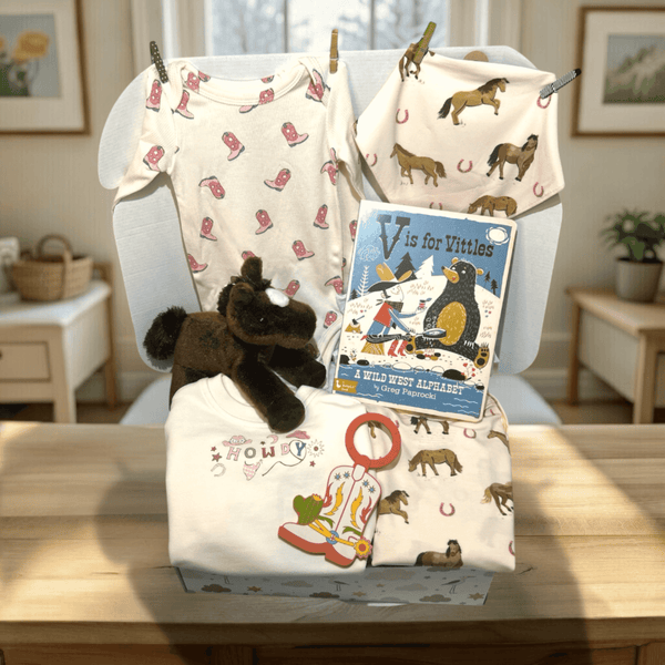 Cowgirl Baby Shower Gift  Baby gift basket with clothes, plush toy, and book on a table in a room setting.