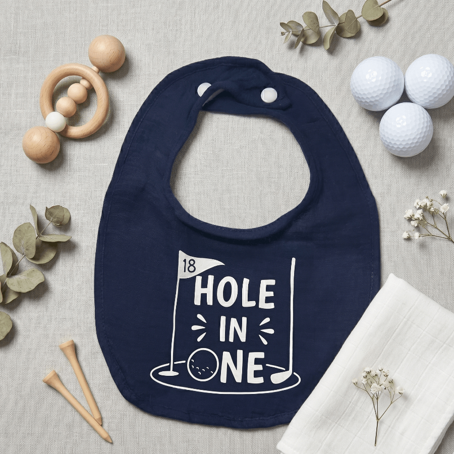 Navy blue bib with '18 Hole in One' print on a neutral background with golf-themed items.
