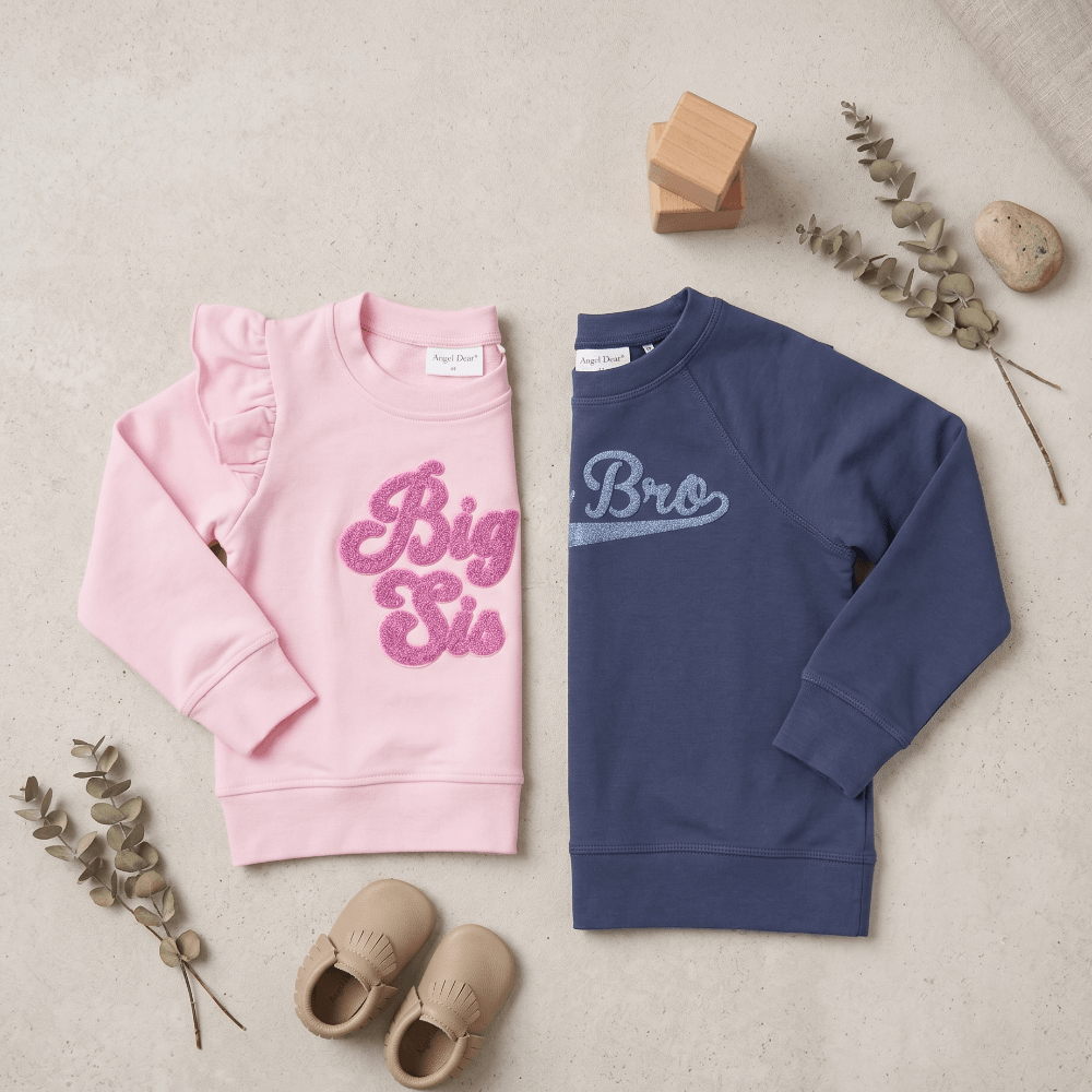 Pink and blue children's sweatshirts with text, accompanied by baby shoes and wooden blocks on a neutral background.