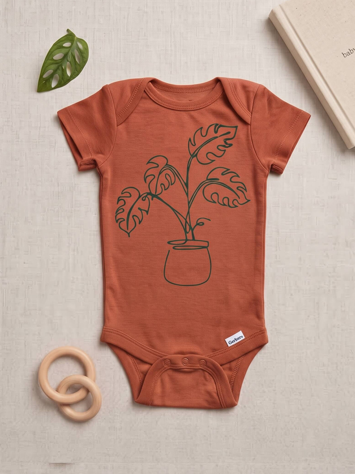 A terracotta-colored baby bodysuit with a black line art graphic of a potted plant, styled in a flat-lay on a textured beige fabric background with a wooden ring toy, a green leaf, and a book.