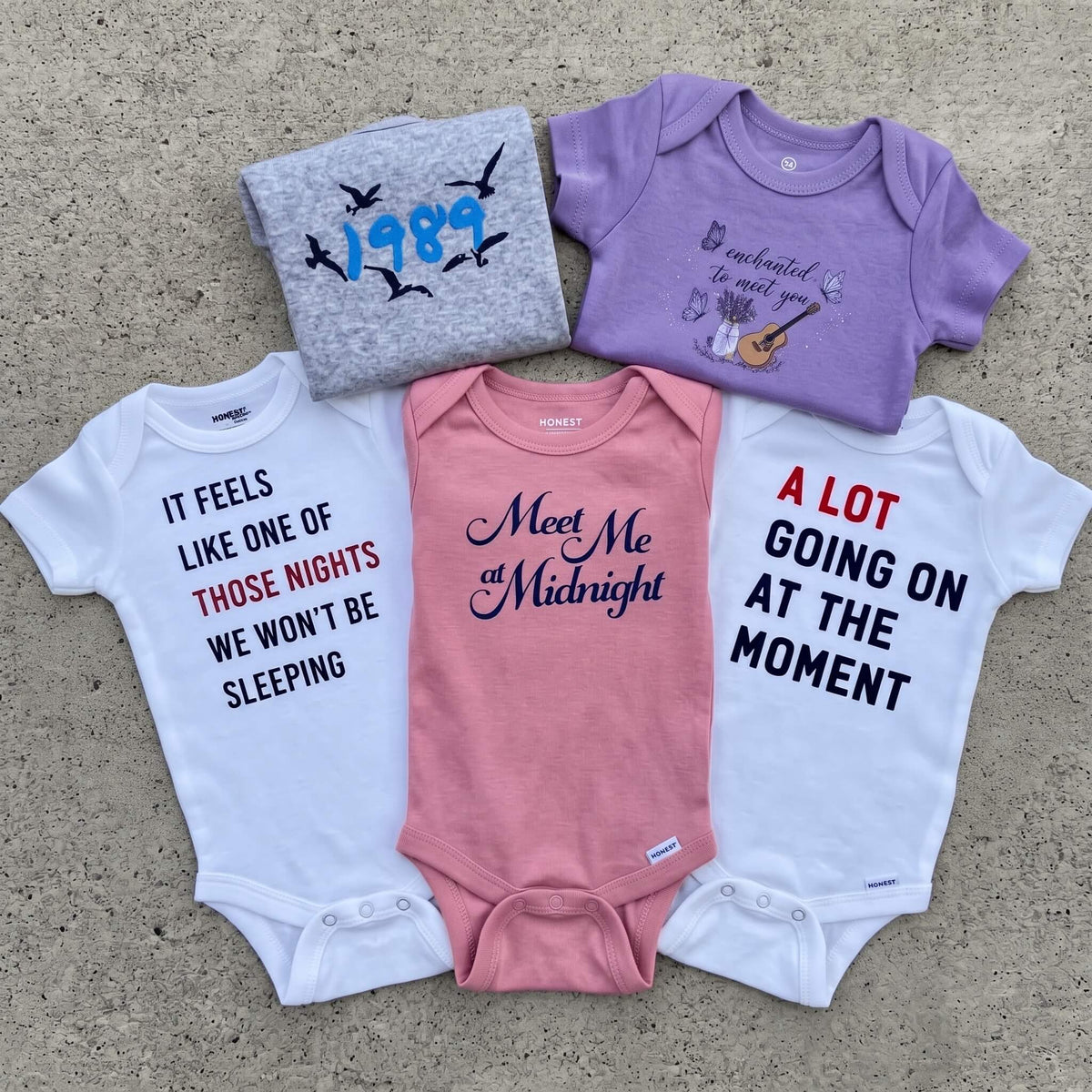 Five baby bodysuits in different colors are arranged on a concrete surface. The set includes a gray sweatshirt-style top with blue '1989' bird graphics, a lavender short-sleeve top with a guitar graphic and text, a white short-sleeve top with black and red text, a pink short-sleeve top with 'Meet Me at Midnight' in navy script, and a white short-sleeve top with black and red text.