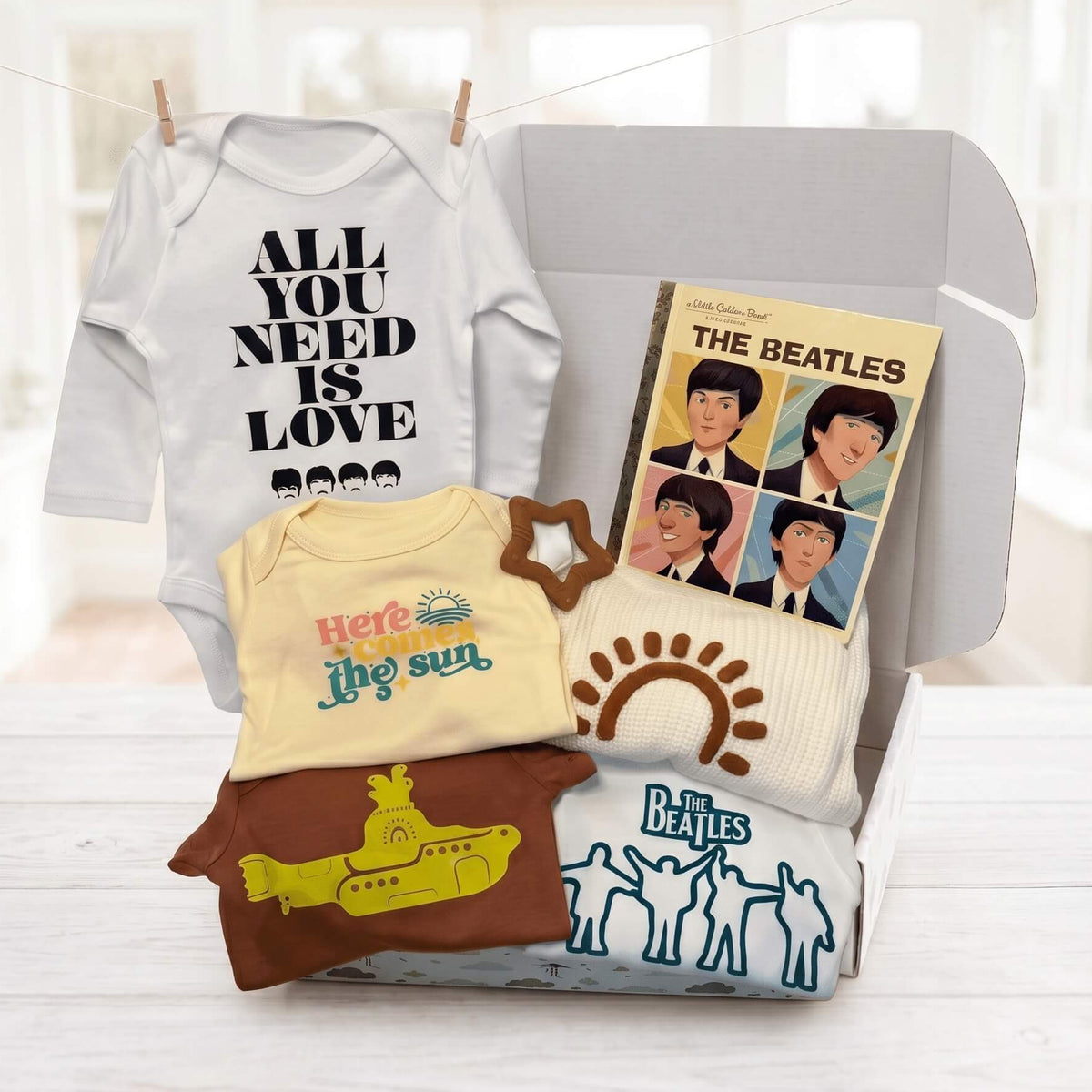 A Beatles-themed baby gift set arranged in an open gray box on a light wood surface, with a white bodysuit reading 'ALL YOU NEED IS LOVE 1968,' a cream shirt with 'Here comes the sun,' a brown T-shirt with a yellow submarine graphic, a cream knit pillow with a brown sun, and a Beatles album cover card.