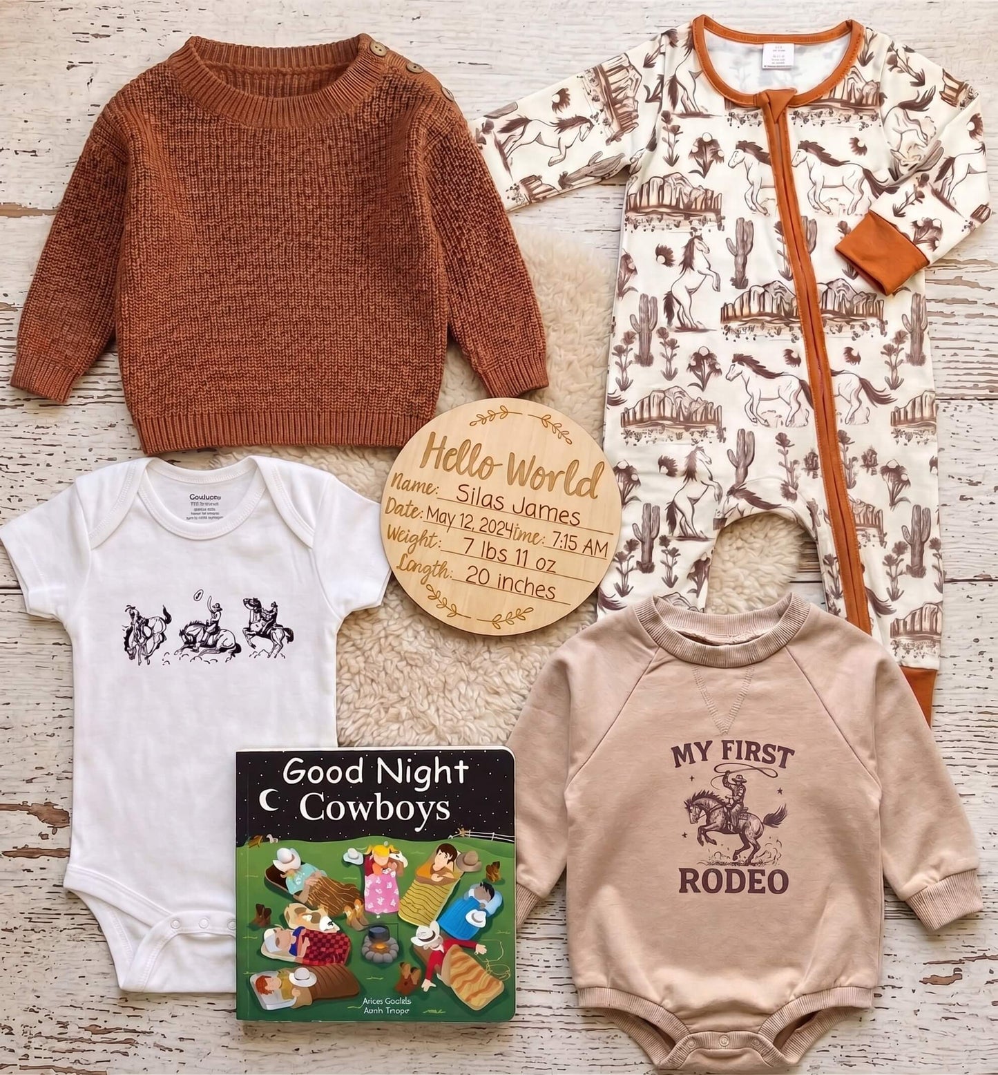 A baby gift set arranged on a whitewashed wood surface, including a rust-brown knit sweater, a cream one-piece with cowboy print, a white bodysuit with a horse-and-rider graphic, a beige long-sleeve shirt with a rodeo graphic, a personalized wooden name card, and a small 'Good Night Cowboys' book.