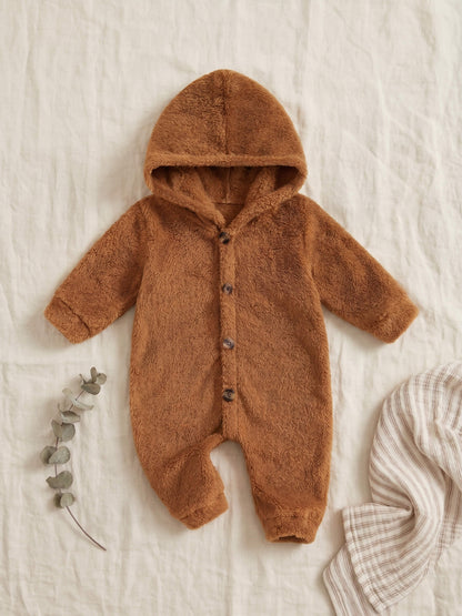 A baby's long-sleeve hooded romper in a soft, fuzzy brown fleece fabric, styled flat-lay on a textured off-white linen background with a sprig of eucalyptus and a striped cloth.