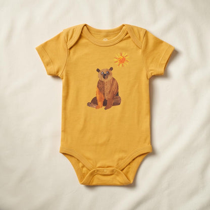 A mustard yellow short-sleeve baby bodysuit laid flat on a white fabric background, featuring a graphic of a brown bear sitting under a small yellow sun.