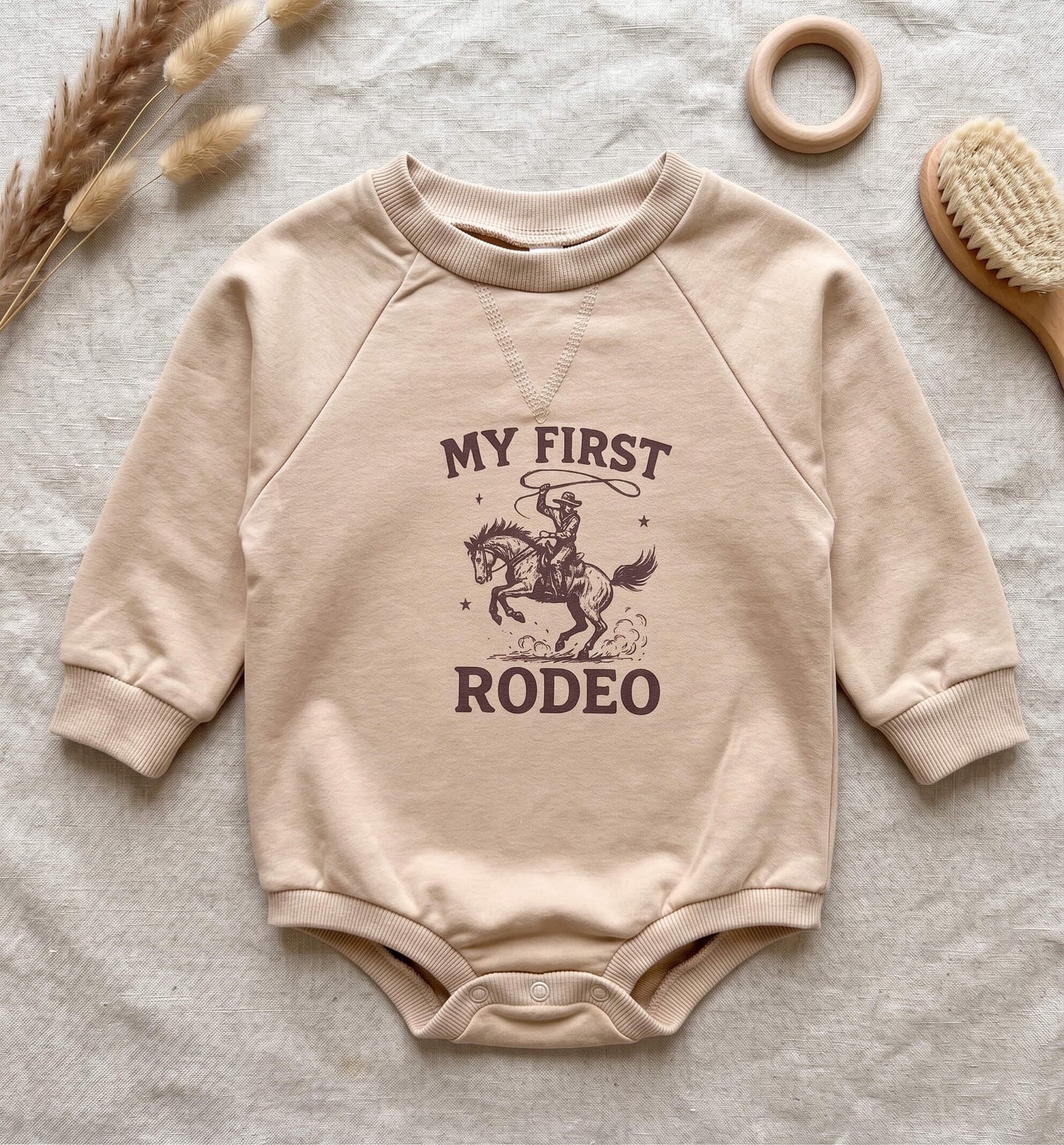 A beige long-sleeve baby bodysuit with a dark brown graphic of a cowboy riding a horse and the text 'MY FIRST RODEO', laid flat on a textured light fabric with decorative dried grass, a wooden ring, and a brush.