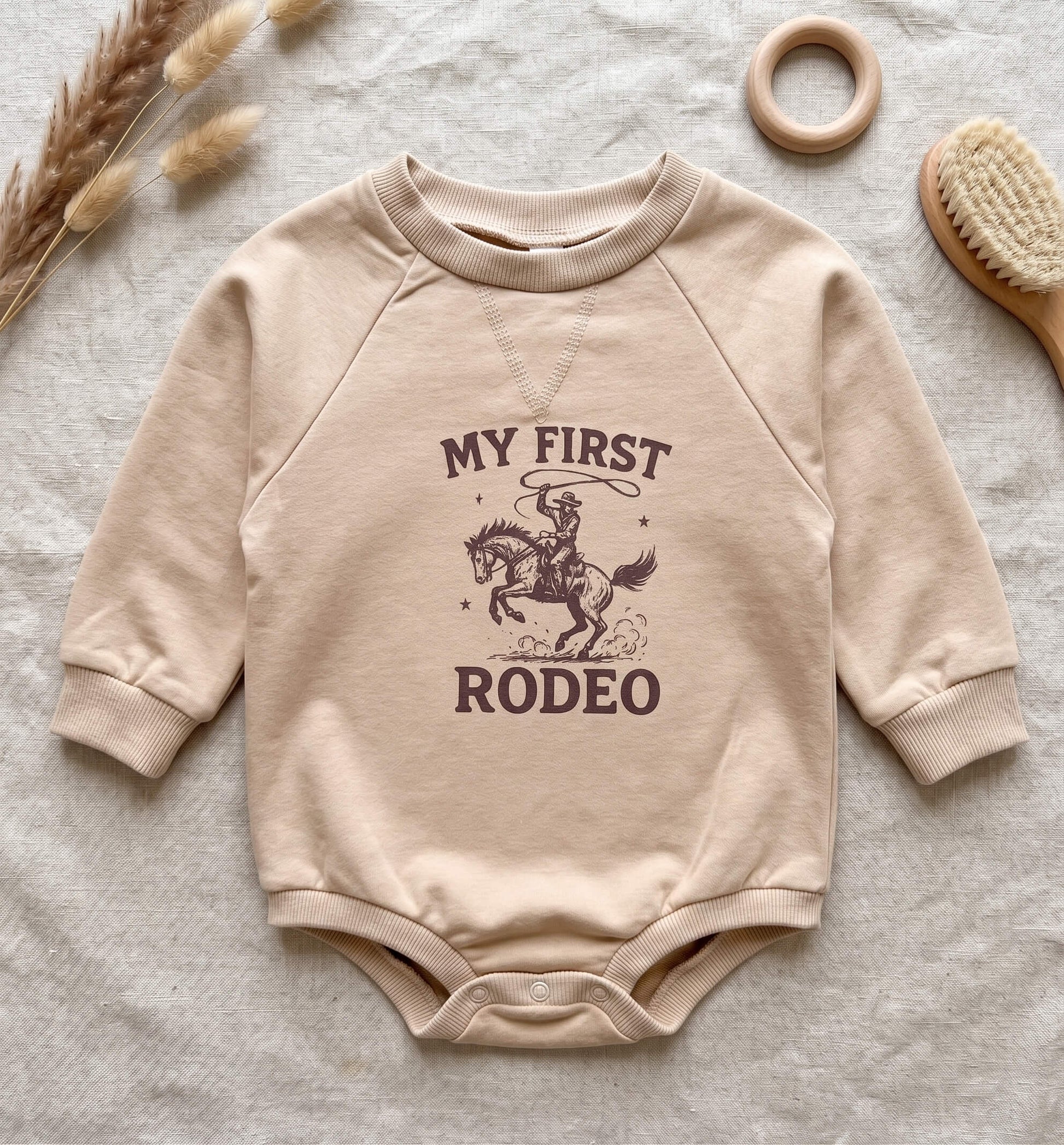 A beige long-sleeve baby bodysuit with a dark brown graphic of a cowboy riding a horse and the text 'MY FIRST RODEO', laid flat on a textured light fabric with decorative dried grass, a wooden ring, and a brush.