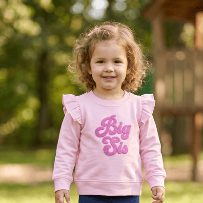 Child wearing a pink 'Big Sis' sweatshirt standing in a grassy area with a playground in the background.