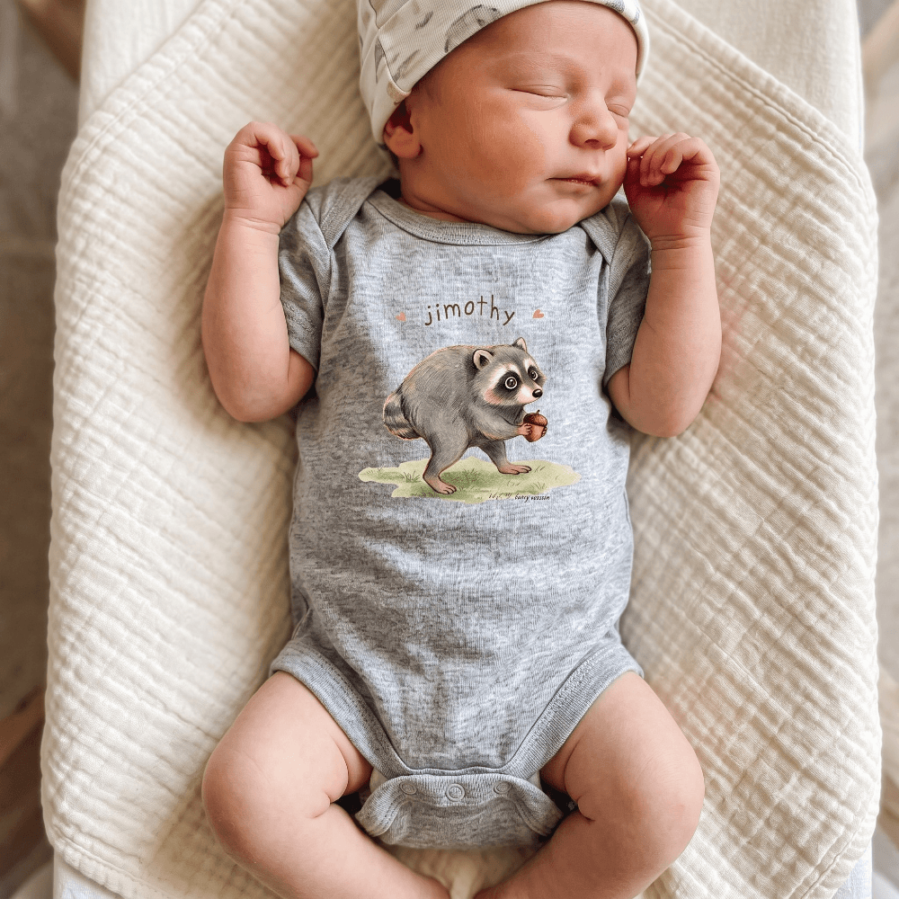 A sleeping newborn baby wearing a heather gray short-sleeve bodysuit with a raccoon graphic and the name 'jimoth' printed on the chest, along with a light patterned beanie, lying on a textured cream blanket.