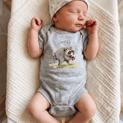 A sleeping newborn baby wearing a heather gray short-sleeve bodysuit with a raccoon graphic and the name 'jimoth' printed on the chest, along with a light patterned beanie, lying on a textured cream blanket.