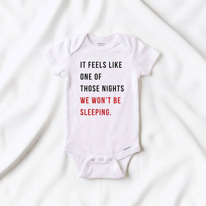 A white short-sleeve baby bodysuit laid flat on white fabric, printed with the text 'IT FEELS LIKE ONE OF THOSE NIGHTS WE WON'T BE SLEEPING.' in black and red lettering.