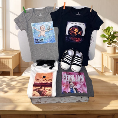 A curated baby clothing set displayed on a chair in a sunlit room. The set includes a heather gray baby bodysuit with a Nirvana graphic, a black baby t-shirt with a Tupac graphic, a white graphic tee with Alice in Chains artwork, a heather gray graphic t-shirt with Pearl Jam artwork, a pair of black-and-white sneakers, and black sunglasses.