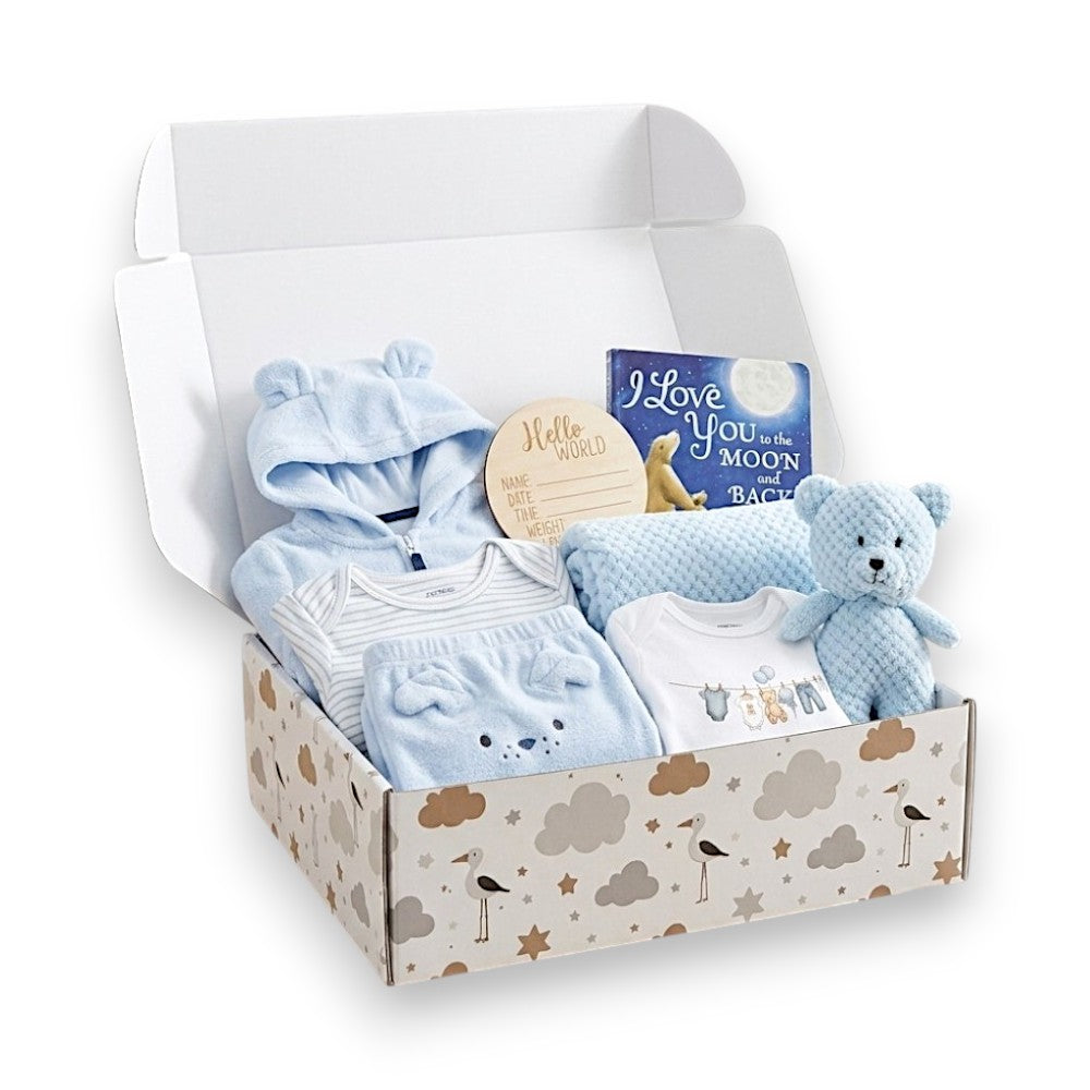 A baby gift set in an open patterned box, featuring light blue baby clothing, a plush teddy bear, a book, and a round wooden keepsake tag on a white background.