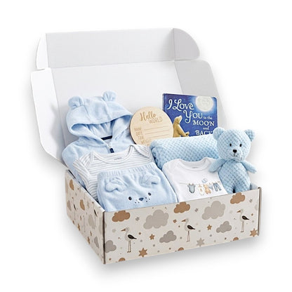 A baby gift set in an open patterned box, featuring light blue baby clothing, a plush teddy bear, a book, and a round wooden keepsake tag on a white background.