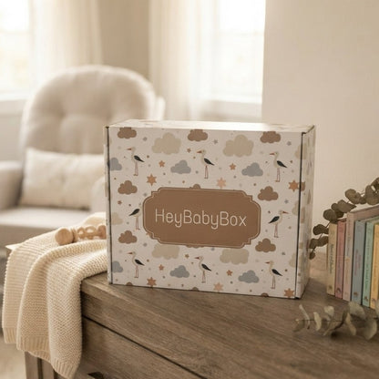 A square product box labeled 'HeyBabyBox' sits on a wooden table in a softly lit room. The box has a cream background with a pattern of clouds, birds, and stars in muted beige, brown, and gray tones.