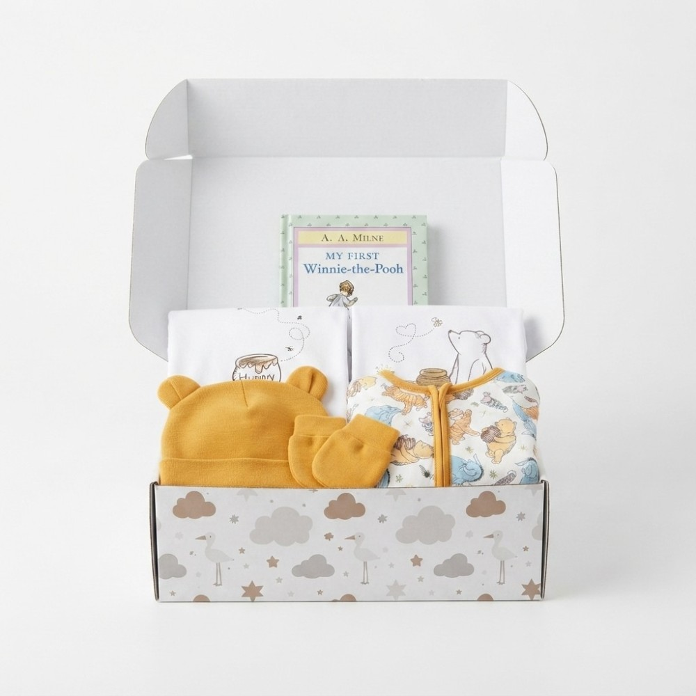 A baby gift set in an open white box, featuring a yellow knit bonnet and matching mittens, a white zippered baby top with Winnie-the-Pod illustrations, folded white towels, and a small book titled 'My First Winnie-the-Pod' by A. A.