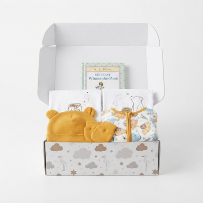 A baby gift set in an open white box, featuring a yellow knit bonnet and matching mittens, a white zippered baby top with Winnie-the-Pod illustrations, folded white towels, and a small book titled 'My First Winnie-the-Pod' by A. A.