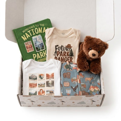 A curated gift box set containing a white baby t-shirt with landscape prints, a beige baby t-shirt with 'Future Park Ranger' text, a blue patterned zippered pouch with outdoor illustrations, a green book titled 'Windows to the National Park', and a brown plush teddy bear, all arranged inside an open patterned gift box.